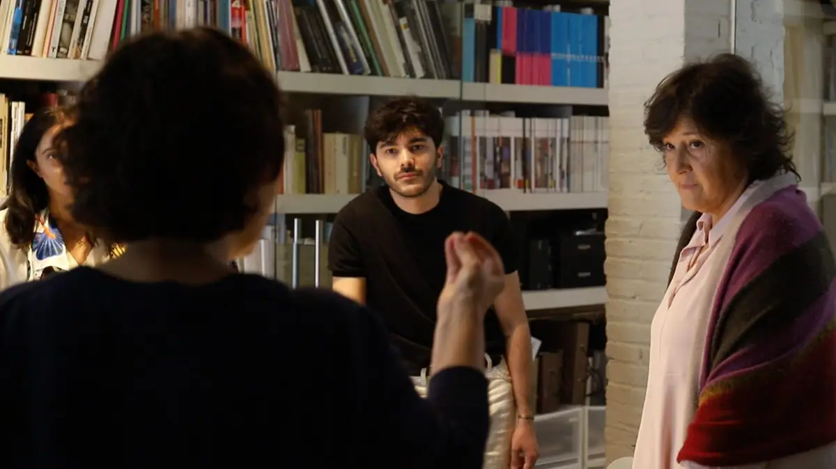 Isabel López Vilalta listens to a colleague explaining something to the team, in front of a bookcase