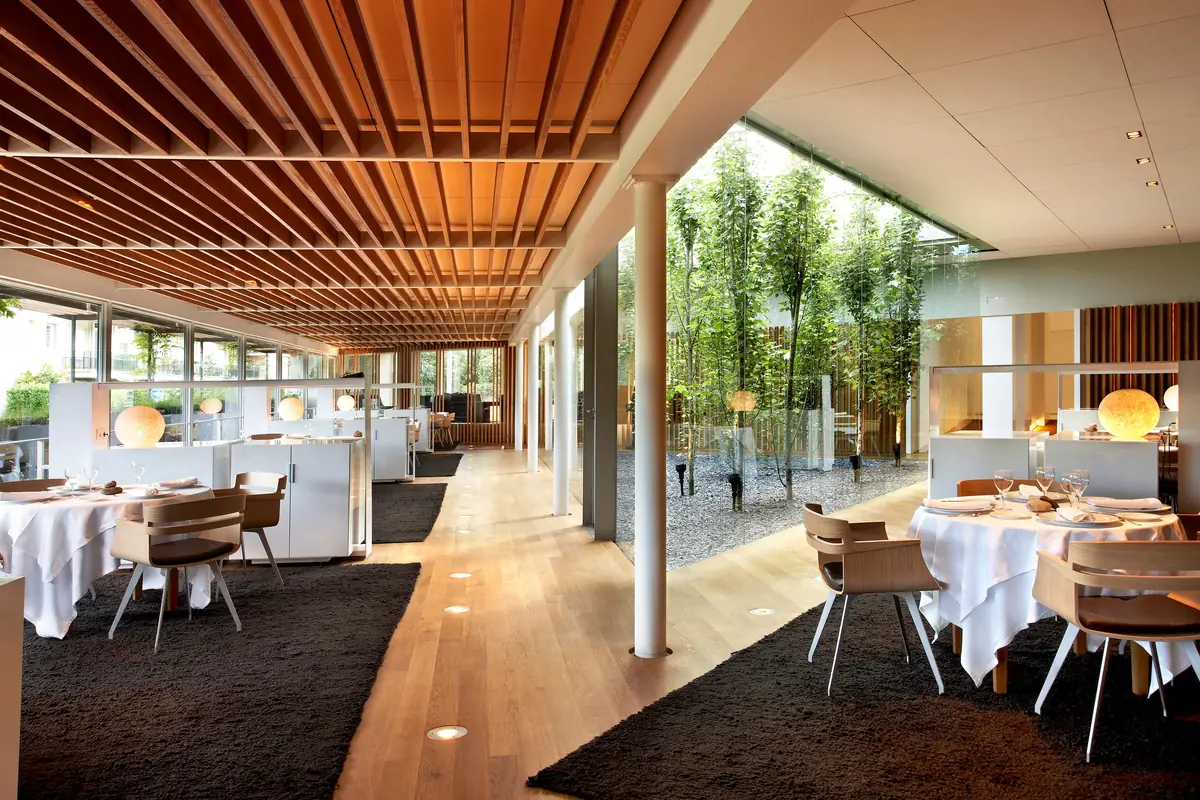 Dining room of El Celler de Can Roca with white-clothed tables, a timber-slatted ceiling and a glazed courtyard with trees