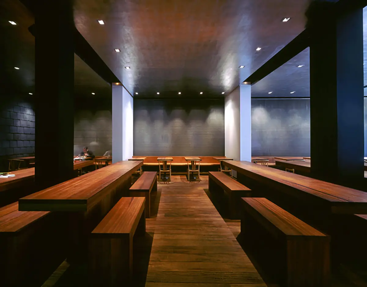 Dining hall of El Japonés with long tables and wooden benches beneath a dark ceiling and grey stone walls