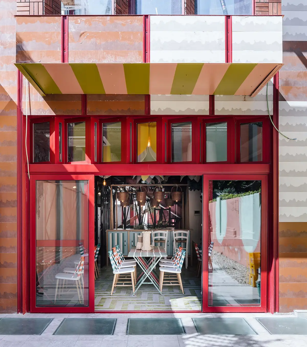 Fachada de tonos terracota con carpinterías rojas y un toldo a rayas; por las puertas abiertas se ve un comedor con lámparas de cesta
