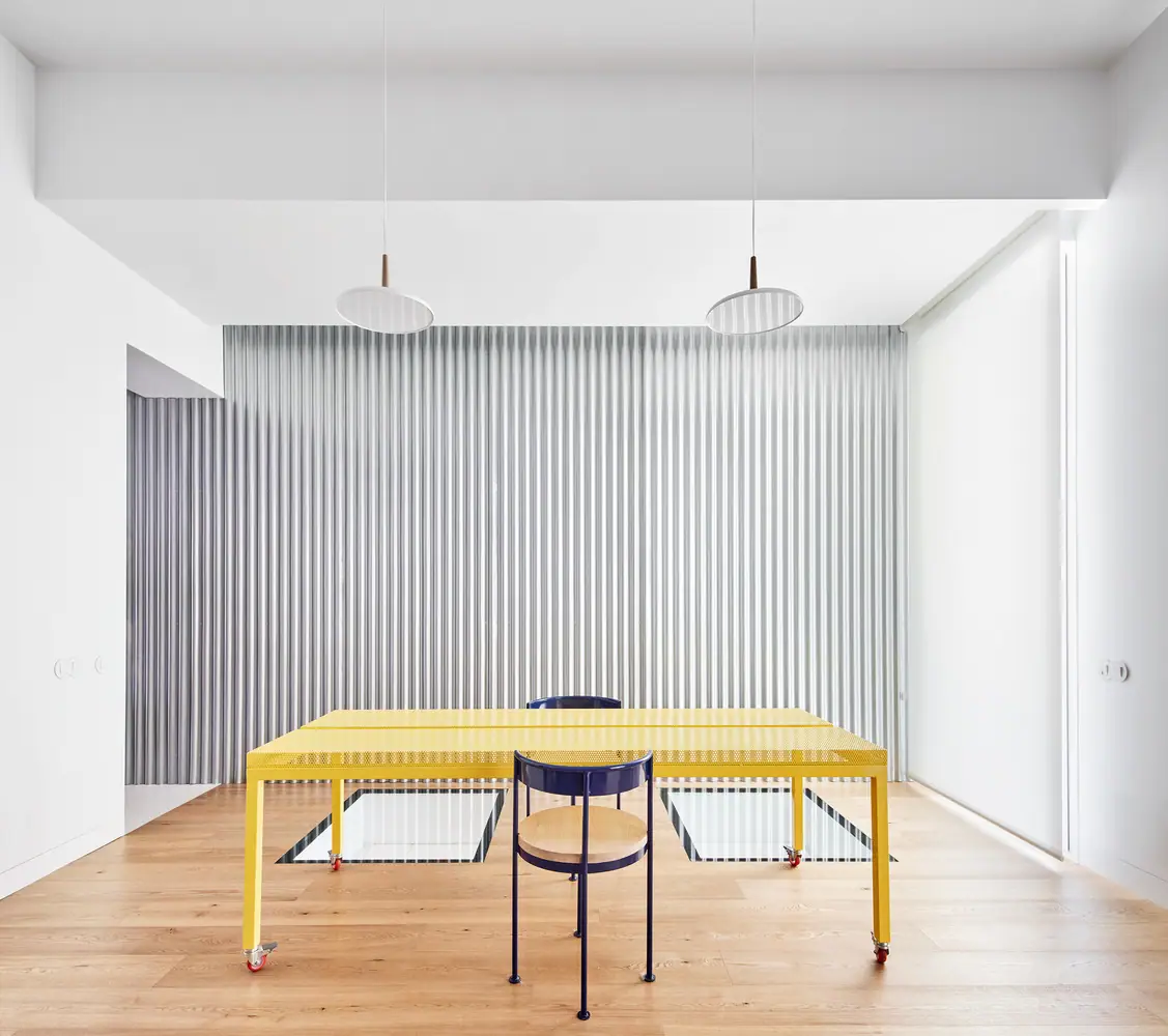 Yellow perforated sheet-metal table on castors and a blue chair in front of a corrugated metal curtain