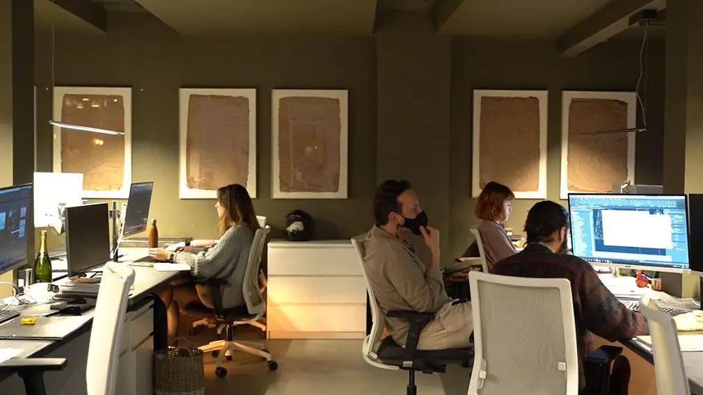 Cuatro personas trabajan ante sus ordenadores en una sala de paredes verde oliva con grandes papeles enmarcados