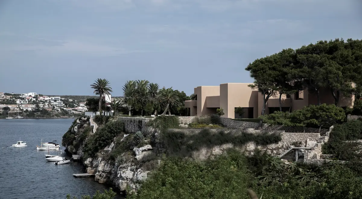 Casa de volúmenes de tono arena entre pinos, sobre un acantilado de la bahía de Mahón con barcas en el agua