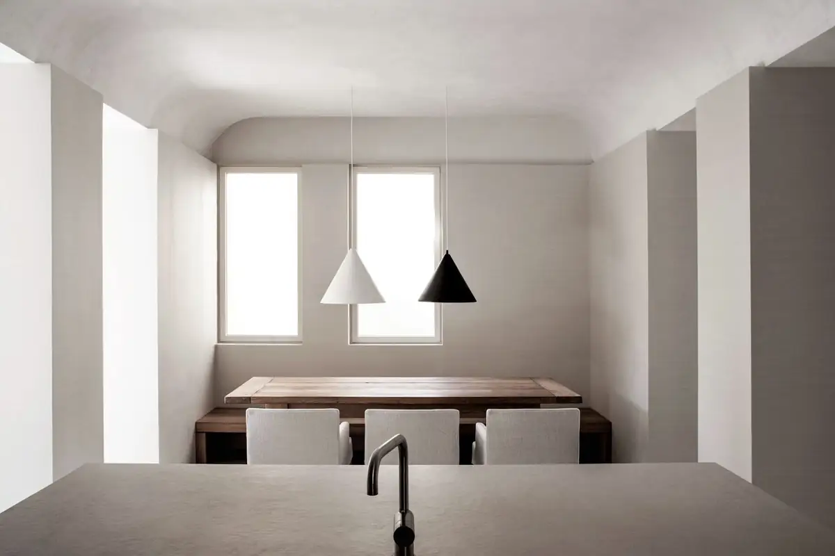 Cocina-comedor blanca con techo abovedado, mesa de madera, sillas tapizadas y dos lámparas cónicas, una blanca y una negra