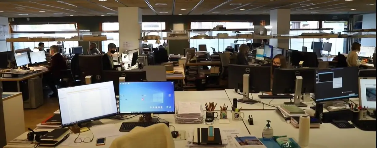 Planta diáfana del estudio con puestos de trabajo, pantallas y maquetas sobre estanterías junto a las ventanas