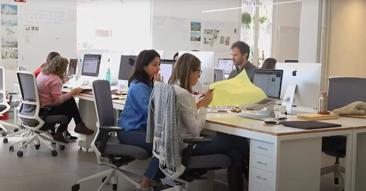 Dos arquitectas revisan un plano junto a sus puestos de trabajo mientras un compañero las observa