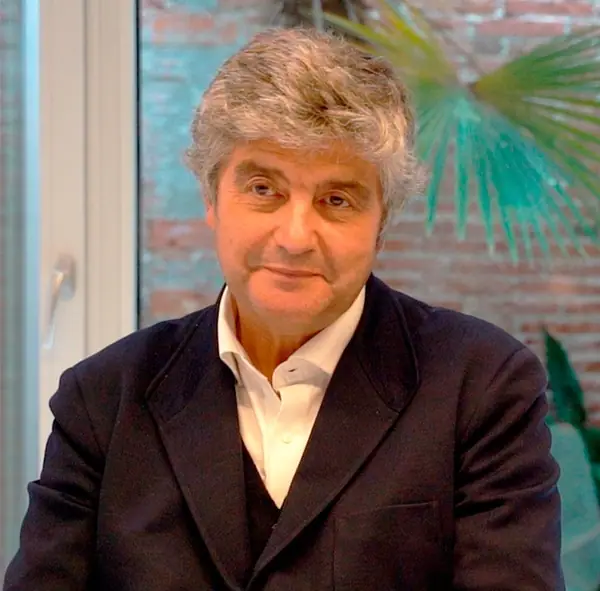 Retrato de Juan Trias de Bes durante la entrevista, con americana oscura y camisa blanca, ante una pared de ladrillo y una palmera