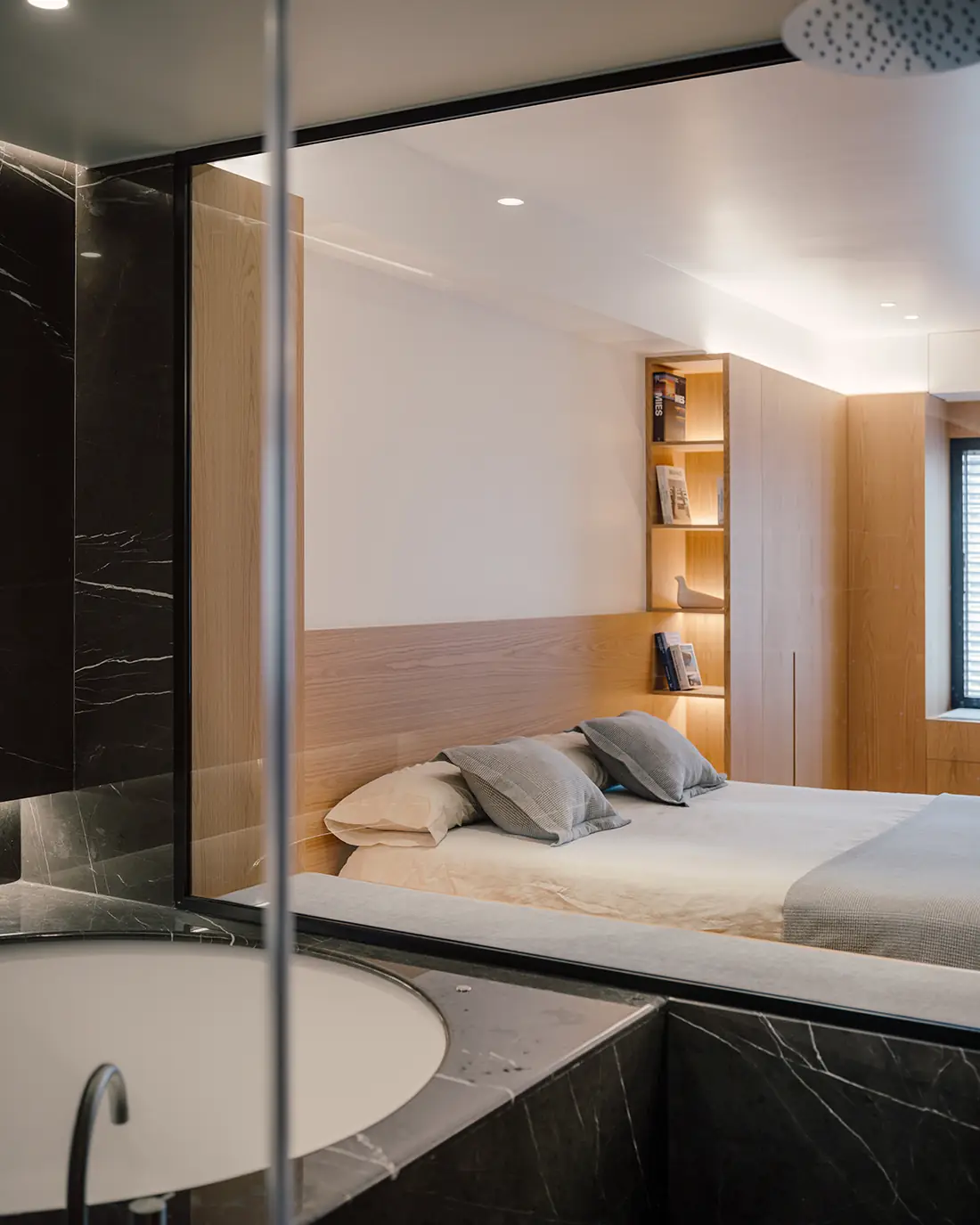 Baño revestido de mármol negro con bañera, abierto mediante un vidrio a un dormitorio con frentes de roble